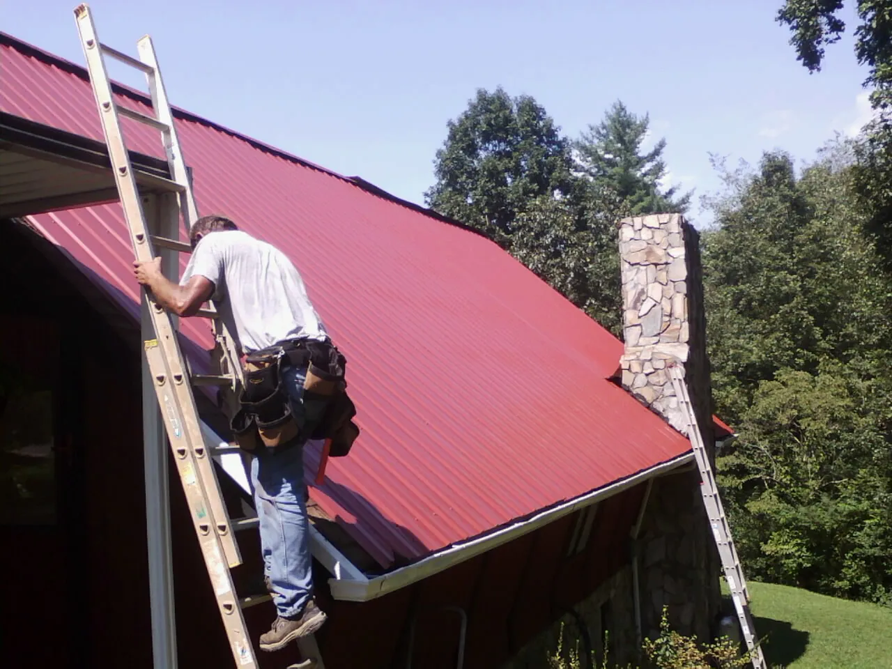 Roofer on a ladder beside a red metal roof and stone chimney — Soffit & Fascia Repair by Roofing Brothers in Yulan